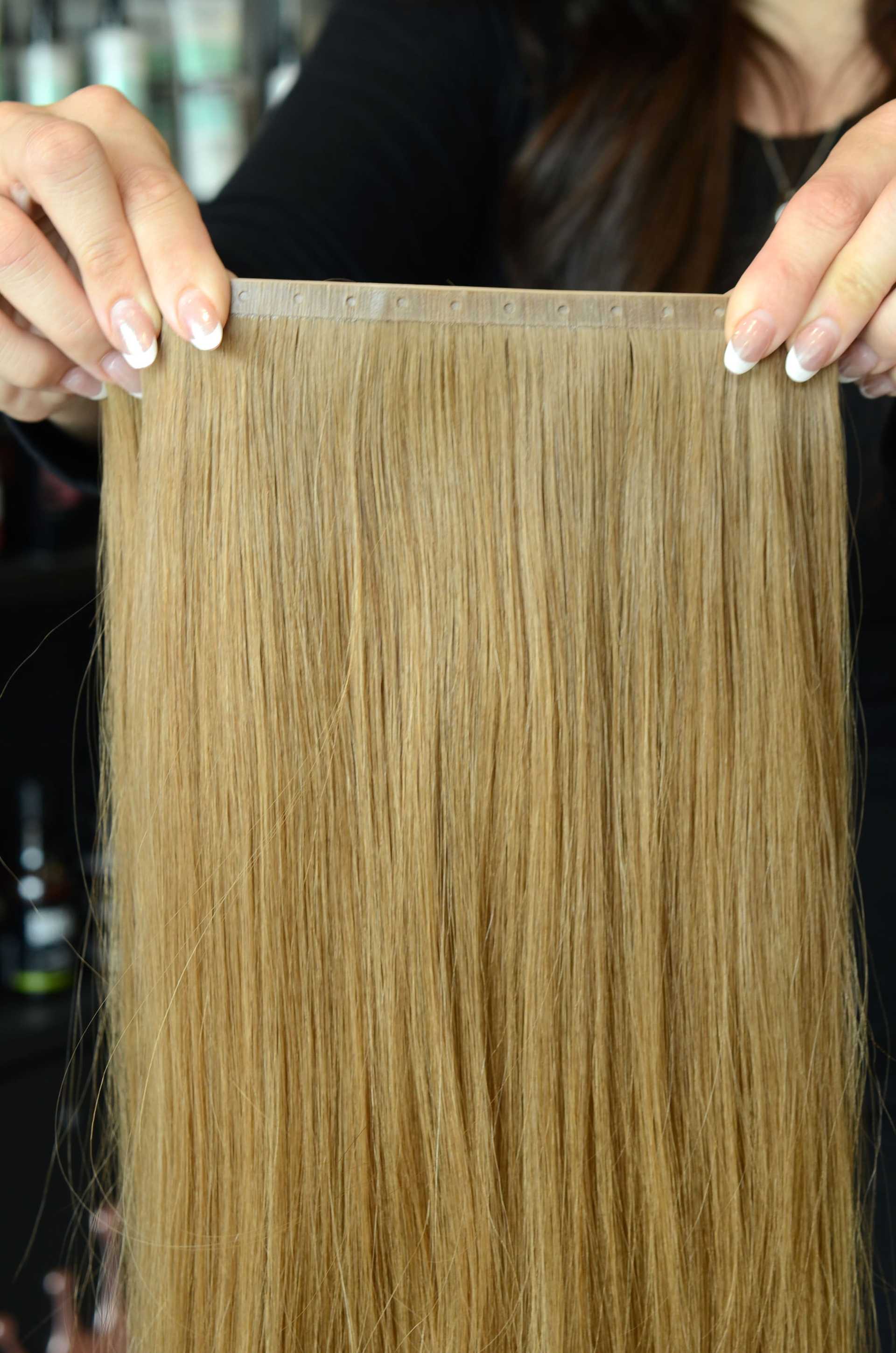 Hands holding a thick, straight blonde hair extension piece.