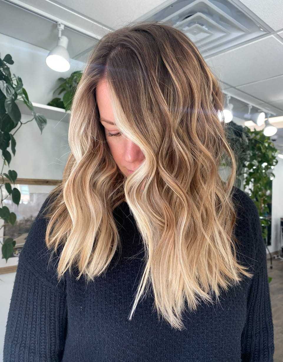 Woman with wavy blonde hair in a black sweater, indoor setting with plants in the background.